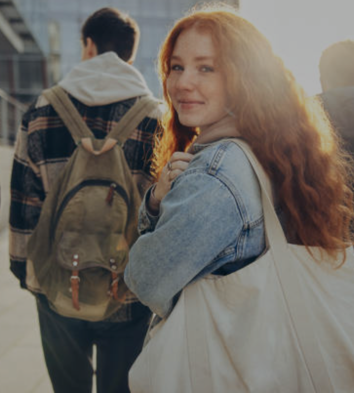 A high school student with red hair smiles over her shoulder while a classmate walks beside her